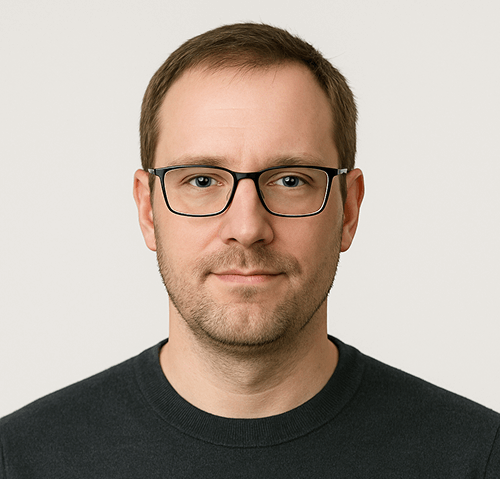 A man with short brown hair, wearing black rectangular glasses and a dark crew-neck shirt, looks directly at the camera against a plain light background.