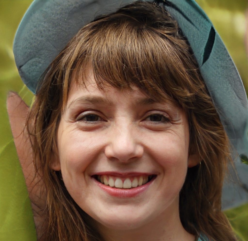 A young woman with light brown hair and bangs smiles warmly at the camera. She is outdoors and wearing a blue hat, with greenery visible in the blurred background.