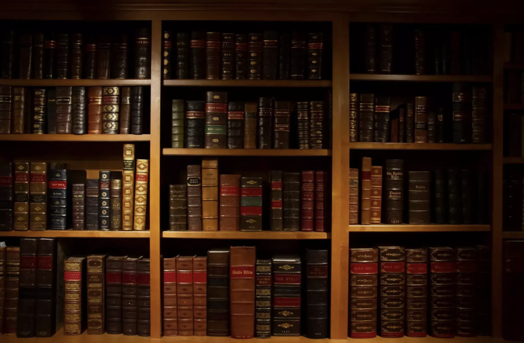 A wooden bookshelf filled with several rows of large, leather-bound books, some with gold lettering on the spines, illuminated warmly from above.