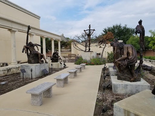 Outdoor sculpture garden with several large, abstract bronze statues on either side of a walkway lined with stone benches. There are plaques near each sculpture and a building with columns in the background.