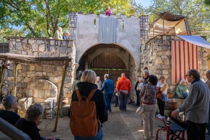 A group of people stand and watch as a person in a pink shirt speaks from atop a stone structure resembling a medieval gate, surrounded by rustic walls and trees under a sunny sky.