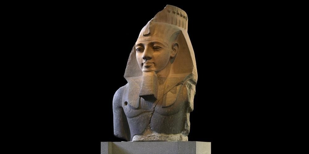 A large stone bust of an ancient Egyptian pharaoh, wearing a nemes headdress with a cobra symbol, false beard, and traditional regalia, displayed against a black background. The sculpture is weathered and partially damaged.