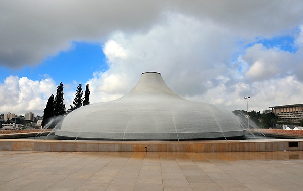 A white, dome-shaped building with water spraying over it, surrounded by a paved courtyard. Trees and modern buildings are visible in the background under a partly cloudy sky.