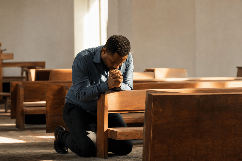 A man kneels and prays with clasped hands at a wooden pew in an empty, sunlit church.
