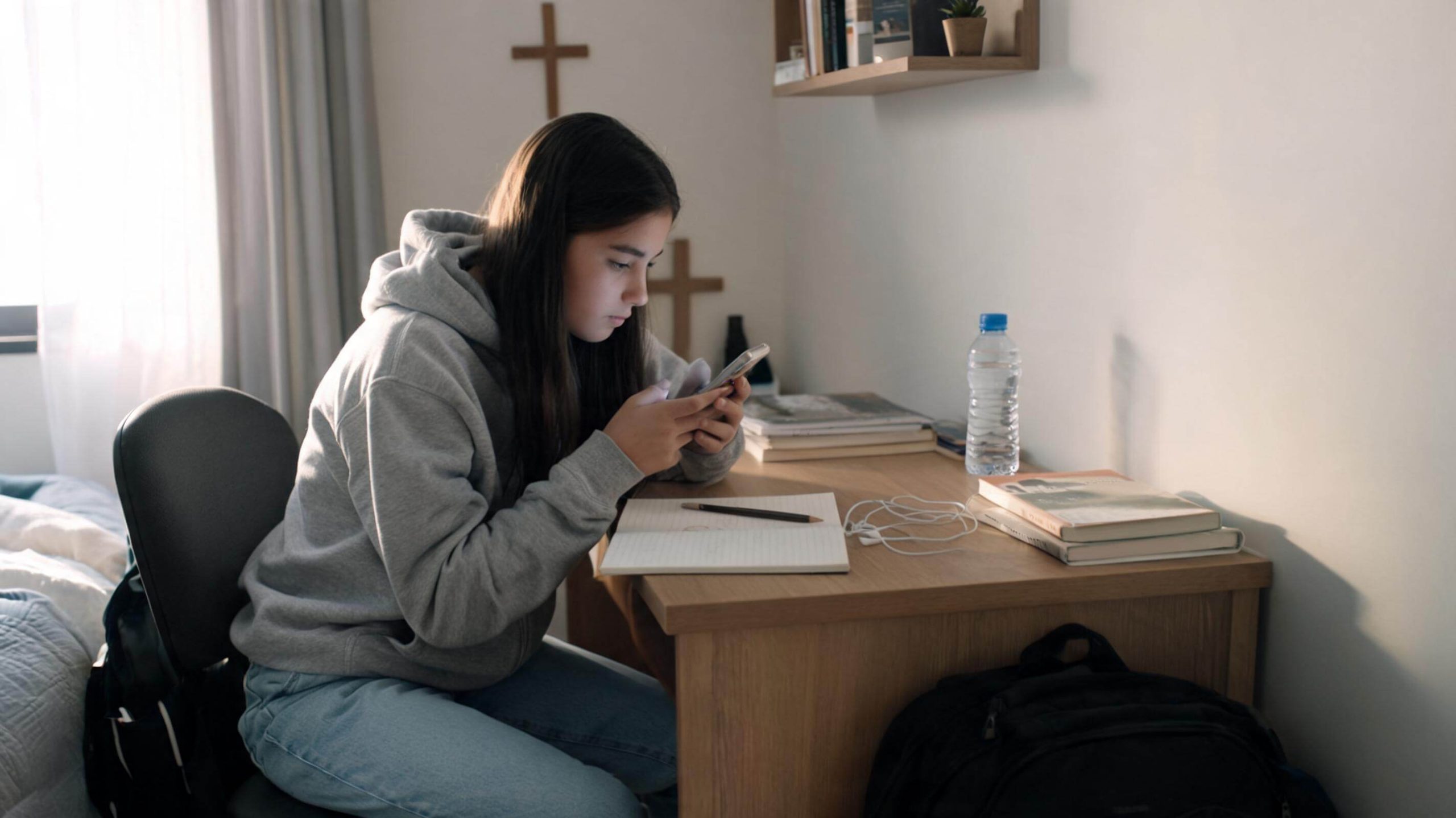 A young woman in a gray hoodie is sitting at a desk, looking at her phone. There are books, a notebook, earphones, and a water bottle on the desk. Crosses are hanging on the wall behind her.