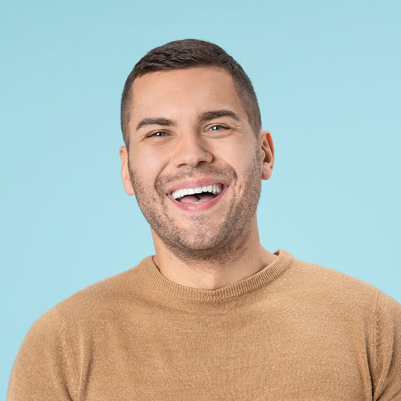 A smiling man with short brown hair wearing a tan sweater stands in front of a solid light blue background.